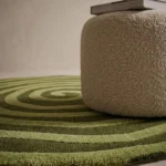 Angle view of the handmade Virel Torelix circular tufted rug showcasing its hypnotic spiral design and green tones next to a boucle pouf.
