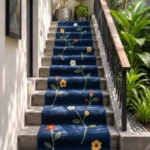 An interior lifestyle photograph of a navy blue floral runner area rug styled elegantly along a stone staircase surrounded by lush green tropical plants.