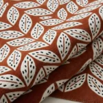 Close-up of the plush tufted pile texture, warm rust-orange background, and intricate black-and-white leaf pattern on the Folivora Leaf rug.