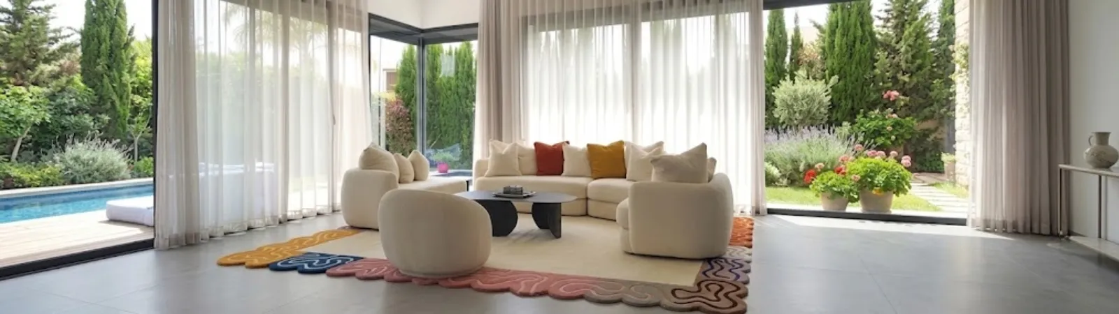 Modern living room interior featuring an organic irregular-shaped colorful bordered area rug anchored beneath a modular sectional sofa and armchairs on a large grey tiled floor.