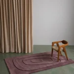 Plumique Basin mauve tufted area rug styled in a contemporary interior space with a wooden armchair and draped curtains.