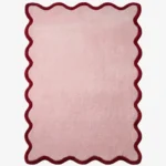 Studio flat-lay product photograph of the rectangular Cheeky Bloom rug on a clean white background, showcasing the solid light pink wool field with a distinct scalloped and wavy dark red border.