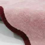 Angled studio product view of the rectangular Cheeky Bloom rug, highlighting its unique scalloped wave edges and plush pink wool surface.